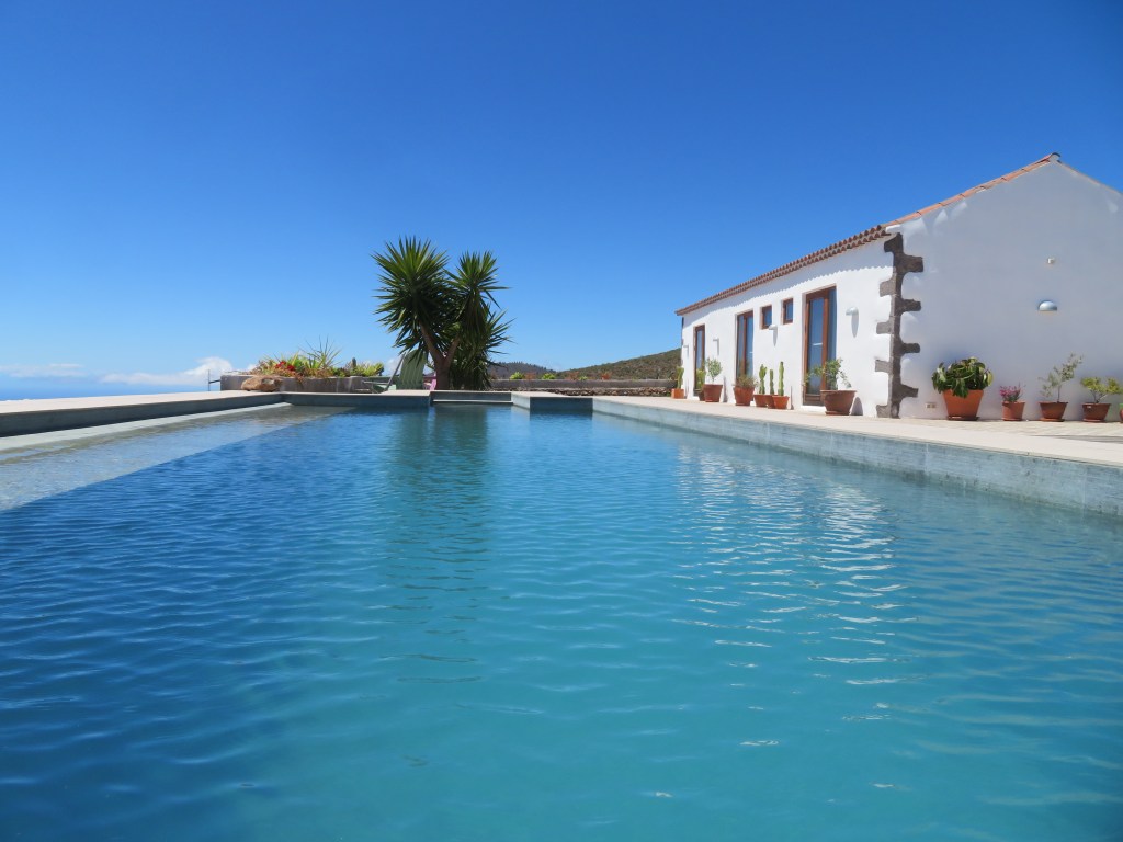 A serene poolside view featuring a clear blue swimming pool, a modern white house with wooden windows, and lush greenery under a bright blue sky.